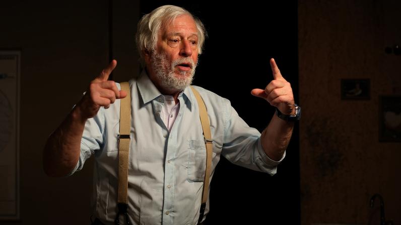 Homem idoso com cabelo e barba brancos, usando camisa azul clara e suspensórios bege, gesticula com as mãos levantadas em cena teatral. Ao fundo, parede amarela com cruz, quadros pequenos e pia com torneira.