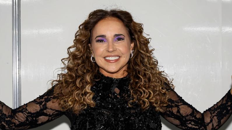 A imagem mostra uma mulher com cabelo cacheado e longo, usando um vestido preto brilhante com detalhes em renda e uma saia curta. Ela está em um ambiente interno, com paredes brancas ao fundo, e faz uma pose sorridente com os braços abertos. A maquiagem inclui sombra roxa e um sorriso expressivo.