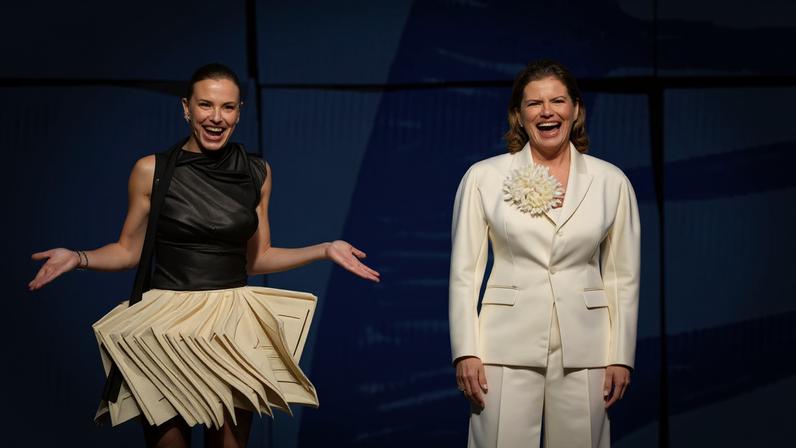 Duas mulheres sorriem em palco com fundo azul. A da esquerda veste vestido preto com saia bege imitando muitas folhas de papel; a da direita usa terno branco acetinado com flor no peito.