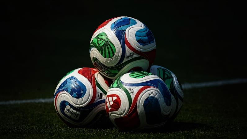 Quatro bolas de futebol oficiais com design colorido em vermelho, azul, verde e branco empilhadas sobre o gramado de um campo. Linha branca do campo visível ao fundo.