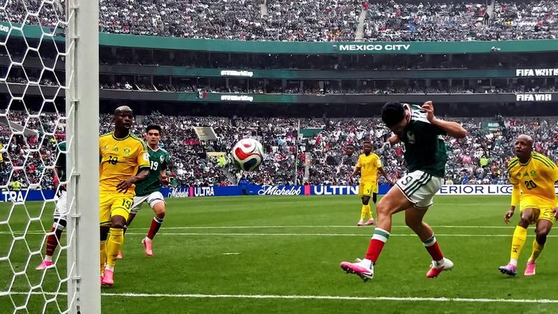 Jogador da seleção mexicana chuta a bola em direção ao gol defendido pelo goleiro jamaicano, com quatro jogadores jamaicanos próximos na área. Estádio cheio com público e faixa indicando 'Mexico City' ao fundo.