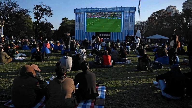 Pessoas sentadas em gramado de parque assistem a jogo de futebol exibido em grande telão. Ambiente ao ar livre ao entardecer com árvores e edifícios ao fundo.