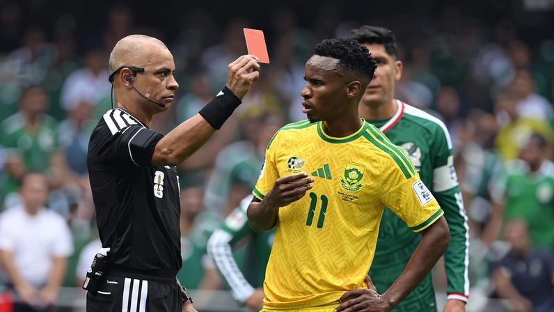 Árbitro com uniforme preto levanta cartão vermelho em direção a jogador da África do Sul com camisa amarela número 11. Jogador reage com a mão no peito e expressão de surpresa. Ao fundo, jogador do México com uniforme verde observa a cena.