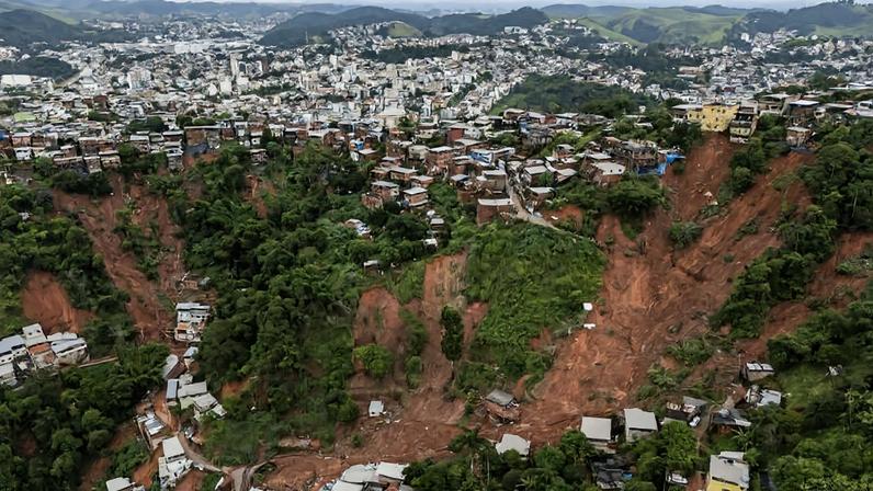 Deslizamento de terra em encosta de morro em área urbana, com grande faixa de terra exposta e casas destruídas ou danificadas. Vegetação ao redor e cidade ao fundo sob céu nublado.