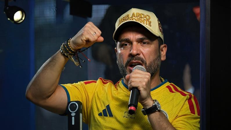 Homem com boné bege e camiseta amarela canta ao microfone, com punho direito erguido em gesto de força. Fundo iluminado em azul e roxo, ambiente de show ou evento musical.