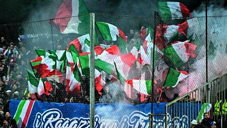 Torcida agitada em arquibancada de estádio, com várias bandeiras italianas verdes, brancas e vermelhas tremulando. Fumaça branca envolve o grupo, que está atrás de uma grade de proteção. Banner azul com texto em italiano está pendurado na frente.