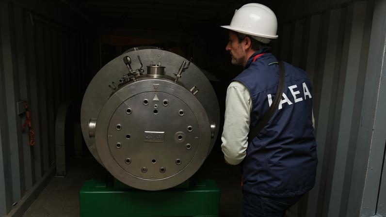 Homem com capacete branco e colete azul com sigla AIEA examina equipamento metálico cilíndrico dentro de contêiner metálico cinza. Equipamento está apoiado em base verde.