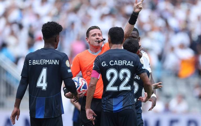 Árbitro de futebol com uniforme laranja levanta o braço direito mostrando cartão vermelho a jogador durante jogo. Dois jogadores de uniforme preto com nomes 'E. Bauermann' e 'G. Cabral' observam a ação, com público desfocado ao fundo.