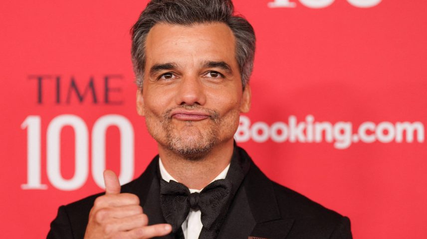 Homem de cabelo grisalho e barba aparada veste smoking preto com gravata borboleta e faz gesto de 'hang loose' com a mão direita. Fundo vermelho com logo TIME 100 e Booking.com visível.