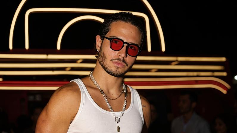 Homem de camiseta regata branca e calça clara usa óculos de sol vermelhos e colar com crucifixo. Ele está em ambiente interno com iluminação neon e pessoas ao fundo. Igor Jansen prepara estreia em 'Por Você' e acumula projetos no cinema