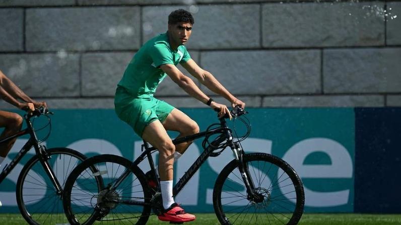 Homem jovem veste uniforme verde e pedala bicicleta preta em campo gramado, com parede de pedra ao fundo.