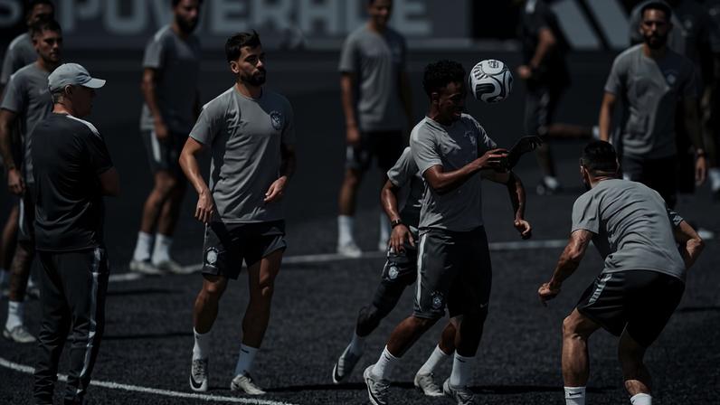 Dez jogadores de futebol vestindo uniformes azuis treinam em campo gramado durante o dia. Um jogador no centro controla a bola com o peito enquanto outros se posicionam ao redor. Um técnico com boné amarelo observa a atividade de perto.