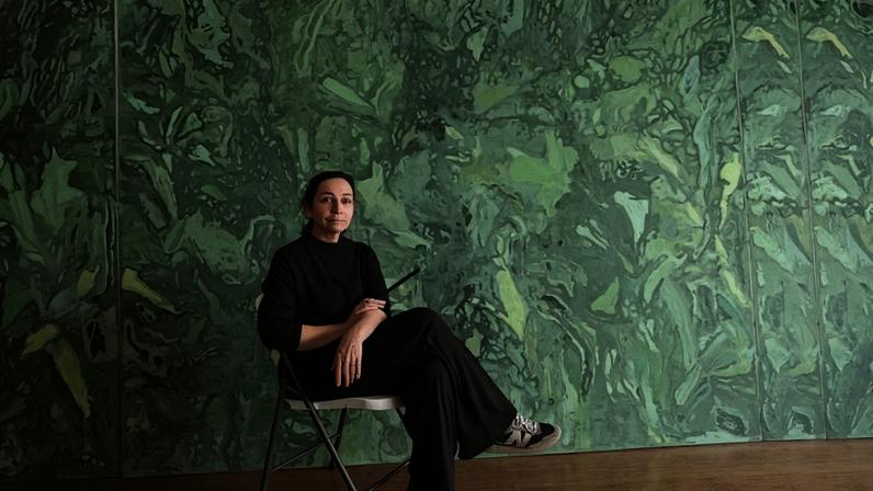 Mulher com roupa preta sentada em cadeira dobrável cinza, com pernas cruzadas, diante de grande pintura abstrata com predominância de verdes, rosas e vermelhos. Piso de madeira clara e parede coberta pela obra vibrante.