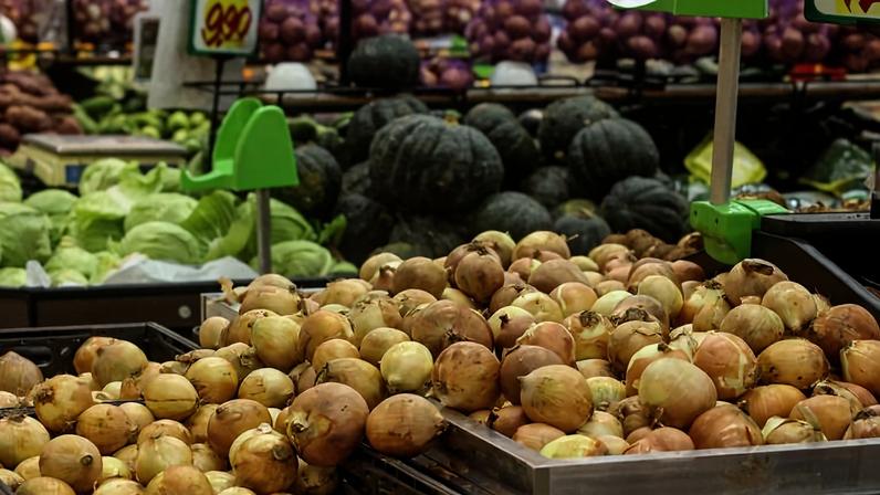 Bancada de feira com cebolas amarelas em primeiro plano. Ao fundo, abóboras verdes, repolhos e outros legumes organizados em caixas.