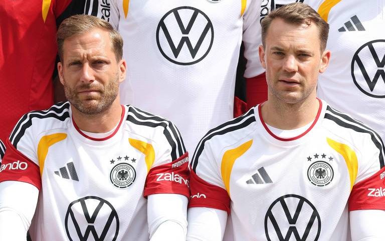 Dois jogadores da seleção alemã vestem uniformes brancos com detalhes em preto, vermelho e amarelo, com o escudo da federação e o logo da Volkswagen no centro. Ambos estão sentados lado a lado, olhando para frente, com expressão neutra.
