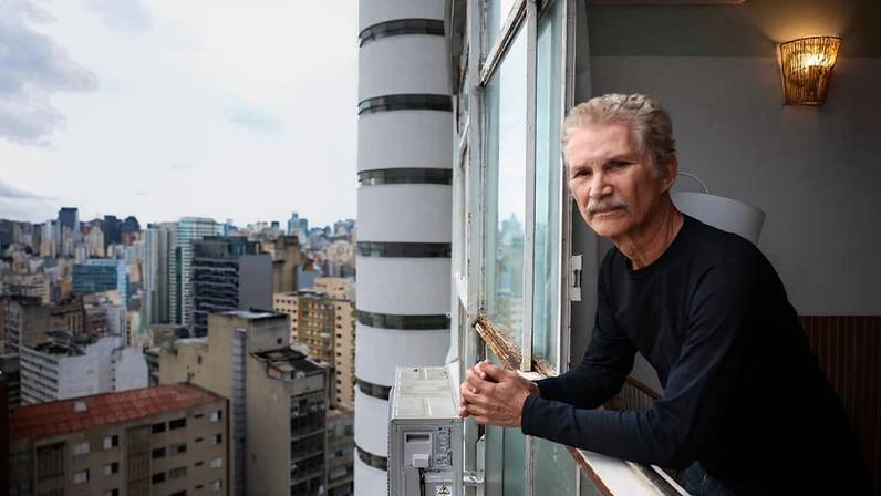 Homem idoso de cabelos grisalhos e camiseta preta apoia os braços na janela de apartamento alto, olhando para a cidade com prédios variados ao fundo em dia nublado.