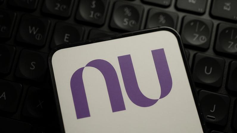Logotipo roxo do Nubank exibido na tela de um smartphone, posicionado sobre um teclado de computador iluminado por luz roxa.