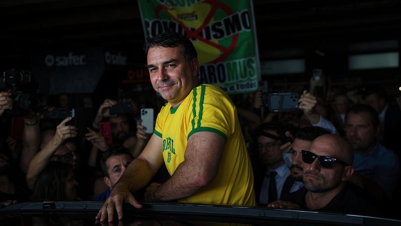 Homem de camiseta amarela com detalhes verdes está parcialmente para fora do teto solar de um carro, cercado por várias pessoas que tiram fotos com celulares e câmeras. Ao fundo, há um cartaz com a palavra 'comunismo' riscada e outras palavras parcialmente visíveis, em ambiente que parece ser uma área coberta, possivelmente um estacionamento ou terminal.