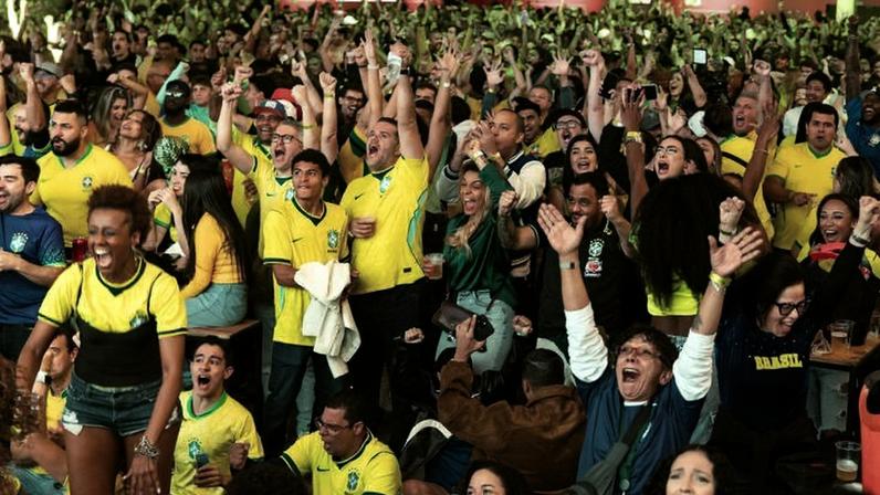 Grande público vestindo camisas amarelas da seleção brasileira, muitos com braços erguidos e expressando entusiasmo em ambiente interno iluminado com luz verde.