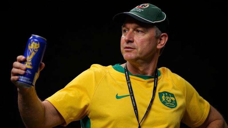 Homem com boné verde e camisa amarela com escudo esportivo segura lata azul com detalhes dourados, em ambiente com público desfocado ao fundo.