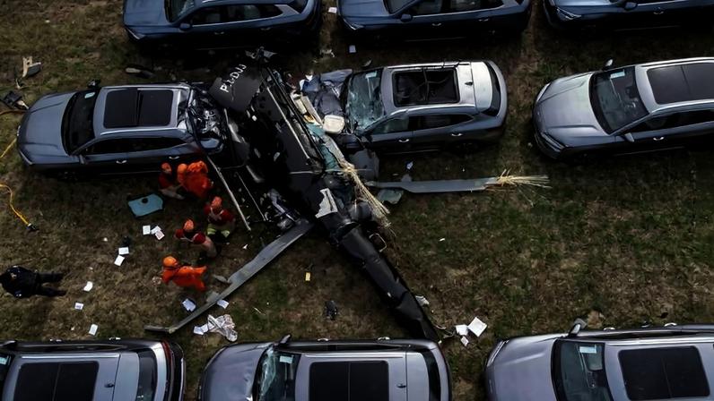 Helicóptero preto da polícia caiu entre carros cinza estacionados em área gramada. Equipe de resgate com uniformes laranja trabalha ao redor da aeronave danificada.