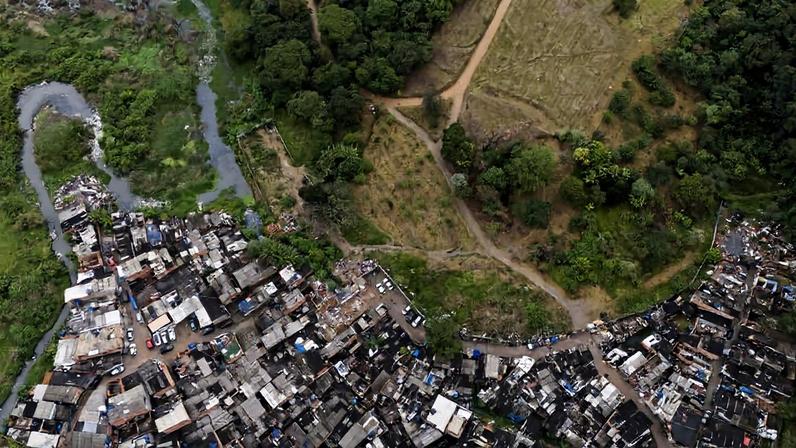 Vista aérea mostra favela com casas de tijolos e telhados variados em primeiro plano, seguida por área verde com árvores e morros ao fundo sob céu nublado.