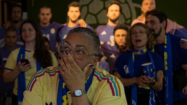 Mulher com óculos e cabelo curto cobre a boca com a mão em gesto de surpresa. Ela veste camisa amarela com detalhes coloridos e cachecol azul e amarelo. Ao fundo, grupo de pessoas também vestindo roupas esportivas, desfocado.