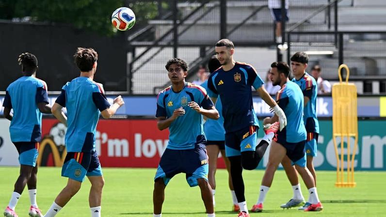 Seis jogadores da seleção argentina de futebol treinam em campo aberto durante o dia. Eles usam uniformes azul e preto com detalhes em amarelo e vermelho, e estão focados em uma bola de futebol que está no ar à frente deles.