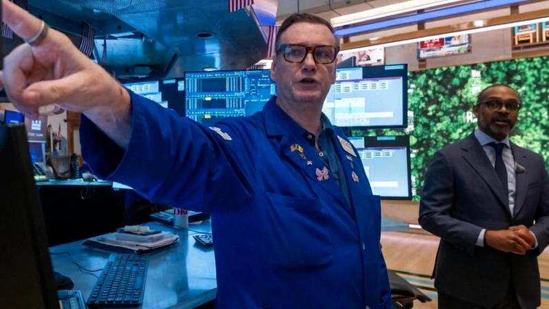 Homem com jaleco azul aponta para um monitor em ambiente de sala de negociação com múltiplas telas e computadores. Outro homem de terno observa ao fundo, com bandeira dos EUA pendurada no teto.