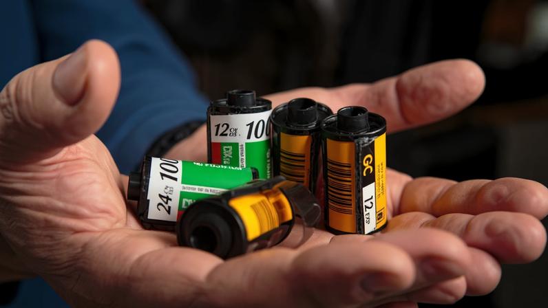 Duas mãos abertas seguram cinco rolos de filme fotográfico analógico, com embalagens em cores verde, branco e amarelo, em ambiente interno com fundo desfocado.