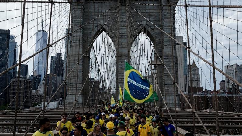 Grupo numeroso de pessoas vestindo amarelo e verde caminha pela Ponte do Brooklyn, em Nova York, carregando uma grande bandeira do Brasil. Ao fundo, arranha-céus da cidade são visíveis sob céu nublado.