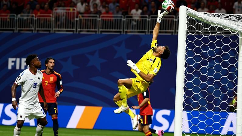 Goleiro vestido de amarelo salta com a mão direita estendida para defender bola próxima ao gol. Jogadores de duas equipes observam a ação, com torcida ao fundo.