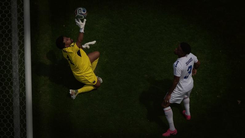 Goleiro vestido de amarelo salta para defender bola próxima ao gol enquanto jogador adversário de branco observa.