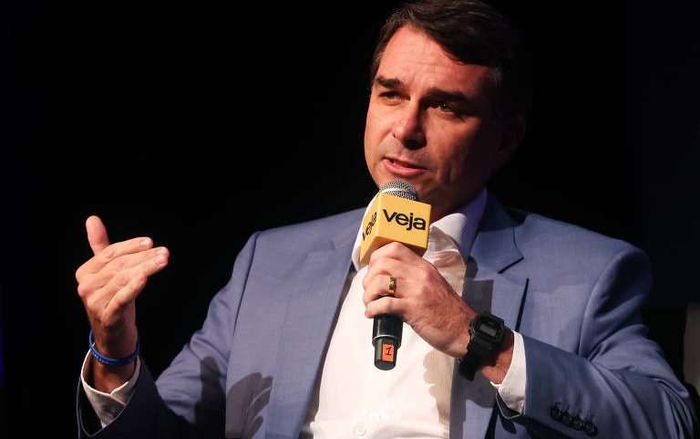 Homem de terno azul e camisa branca segura microfone amarelo com logo da Veja enquanto fala, gesticulando com a mão esquerda. Fundo escuro destaca a figura em primeiro plano.