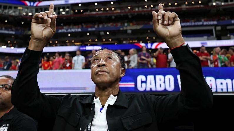 Homem de meia-idade, com jaqueta preta e camiseta branca, levanta os dois dedos indicadores para cima em estádio cheio. Pessoas e placas desfocadas aparecem ao fundo, indicando ambiente de evento esportivo.