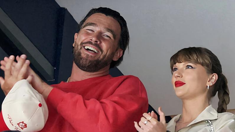 Homem de camiseta vermelha aplaude sorrindo, segurando boné branco, ao lado de mulher de crasaco claro com batom vermelho também aplaudindo. Outras pessoas estão parcialmente visíveis ao redor.