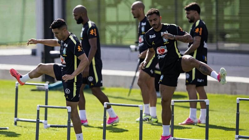 Cinco jogadores de futebol em campo gramado realizam exercícios de aquecimento pulando barreiras baixas. Dois jogadores à frente levantam as pernas sobre as barreiras, enquanto os outros três caminham ao fundo. Todos vestem uniformes pretos com detalhes amarelos e tênis esportivos coloridos.