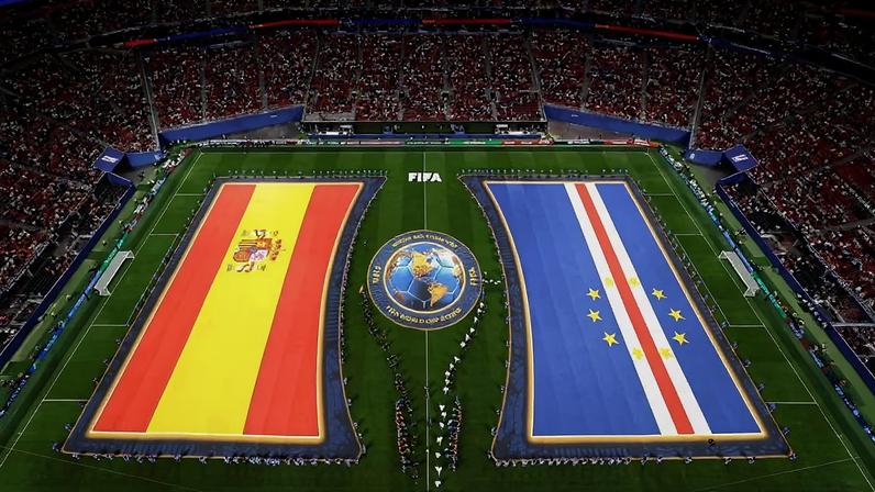 Vista aérea de estádio cheio com torcedores nas arquibancadas. No campo, duas grandes bandeiras da Espanha e da Costa Rica são exibidas por pessoas alinhadas. Telões mostram a taça da Copa do Mundo e imagens relacionadas ao evento.
