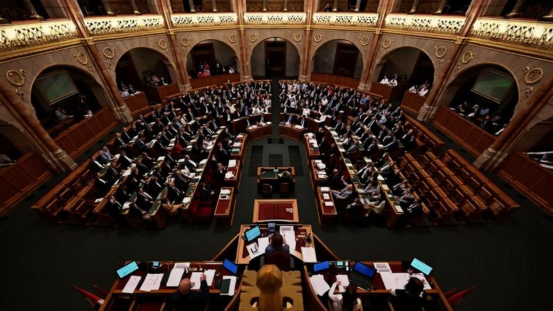 O Parlamento com dezenas de parlamentares sentados em semicírculo em bancos de madeira, em salão com arquitetura clássica, arcos e detalhes ornamentais. Mesa diretora com cinco pessoas em primeiro plano, várias telas e documentos sobre as mesas.
