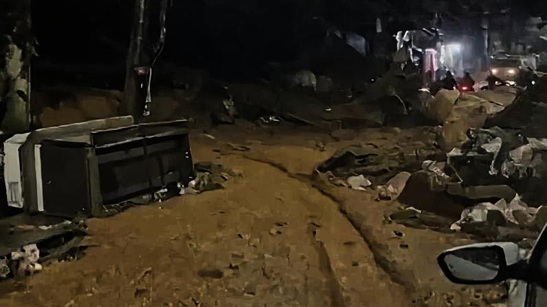 Rua residencial coberta por lama e detritos após deslizamento. Veículo branco estacionado à direita e iluminação pública acesa.