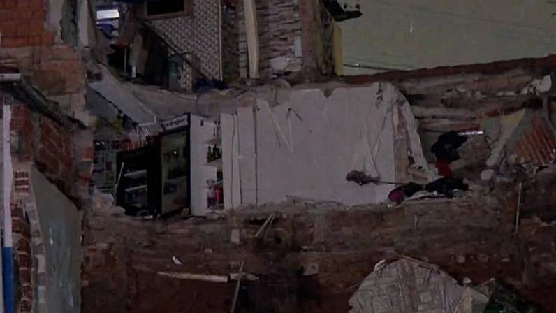 Casa parcialmente desabada em área urbana durante a noite, com escombros visíveis e estrutura danificada. Edifícios vizinhos permanecem intactos, e a cena está iluminada por luz artificial.
