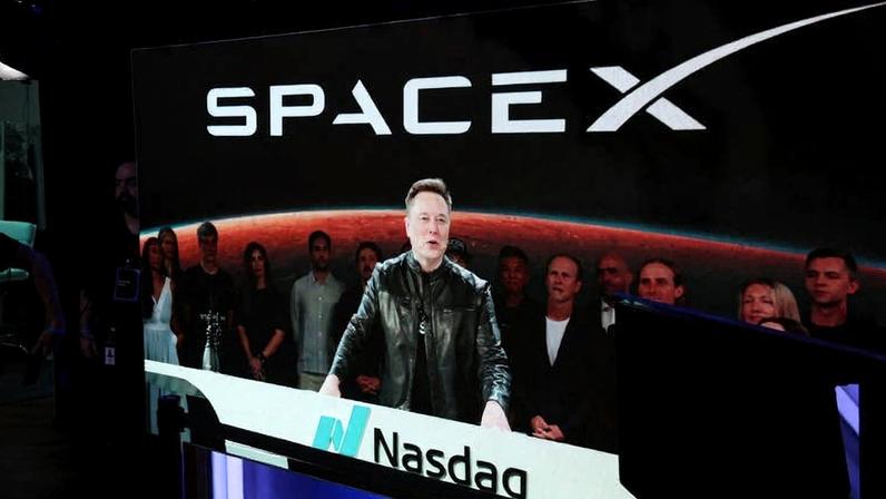 Elon Musk em jaqueta preta fala em púlpito com logo Nasdaq, com painel ao fundo mostrando logo SpaceX e imagem da Terra. Pessoas observam atrás dele.