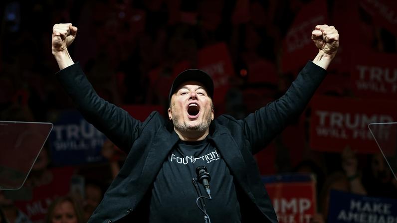 Homem de camiseta preta e boné levanta os braços com punhos cerrados em palanque diante de multidão com cartazes vermelhos e azuis em comício. Elon Musk comemora após discursar em campanha eleitoral de Donald Trump em 2024