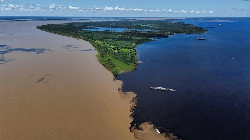 Vista aérea mostra a confluência do Rio Negro de água escura com o Rio Solimões de água barrenta, separados por uma linha nítida ao redor de uma ilha verde.