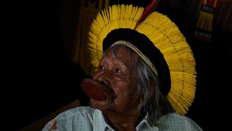 Homem indígena idoso usa cocar amarelo com penas vermelhas e labret grande de madeira na boca. Ele veste camisa branca e está cercado por outras pessoas com roupas tradicionais.