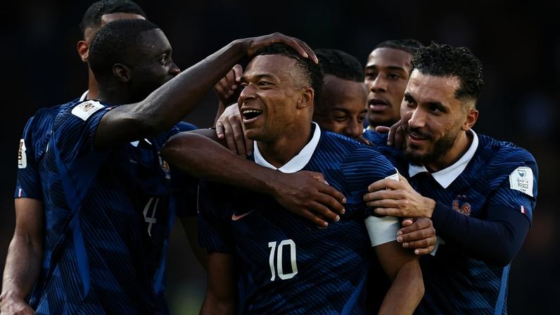 Cinco jogadores da seleção francesa de futebol vestindo uniforme azul escuro comemoram juntos em campo durante partida. O jogador com a camisa 10 está no centro, sorrindo, enquanto os companheiros o abraçam e tocam sua cabeça. O fundo mostra público desfocado nas arquibancadas.