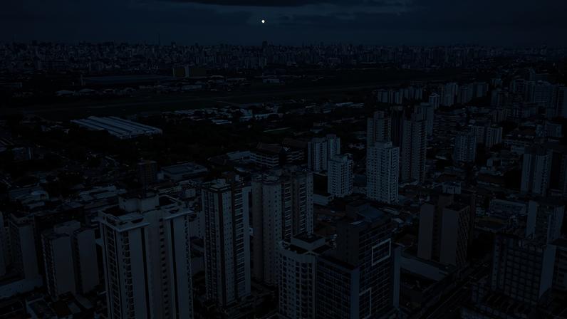 Vista aérea mostra prédios residenciais e comerciais altos em primeiro plano, com pista de aeroporto e áreas verdes ao fundo sob céu nublado.