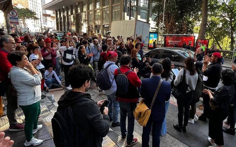 Cerca de 40 pessoas estão reunidas em calçada de área urbana, algumas com mochilas e celulares, observando ou participando de conversa. Ao fundo, edifícios, árvores e veículos em via movimentada.