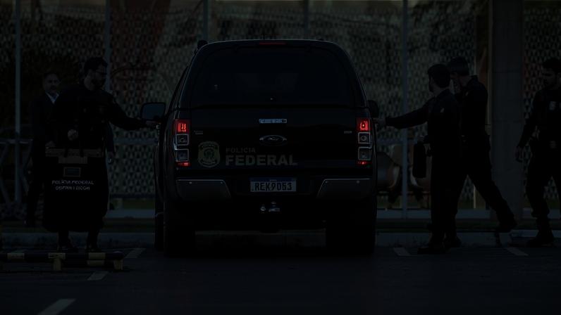 Cinco agentes da Polícia Federal em uniforme preto estão posicionados ao redor de uma caminhonete preta com identificação da PF estacionada em frente a um prédio com fachada de vidro. Um dos agentes segura uma mala preta com a inscrição 'Polícia Federal - Perícia Forense'.