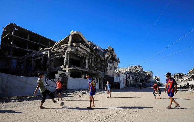 Crianças palestinas jogam futebol em rua da Cidade de Gaza, ao lado de prédios destruídos por bombardeios de Israel